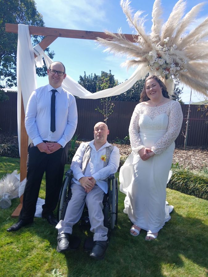 Gray Ruffell, smiling, with a bride and groom under their wedding arch