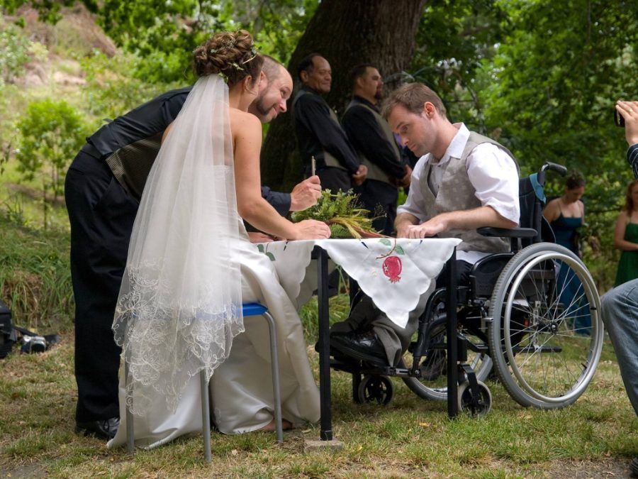 A couple signing their marriage papers at an outdoor table beside Gray